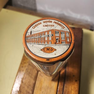 Puede incluir: Un tarro de vidrio vintage con una tapa naranja y blanca. La tapa tiene un gráfico de un edificio con el texto "Canada Motor Products Limited" y "CMP" en un círculo.