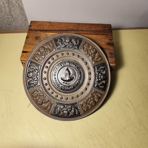 May include: A circular metal decorative plate featuring a central emblem of the Indian Institute of Technology Madras with a lamp icon. The border displays alternating dark and light relief panels with intricate floral and scrollwork patterns.