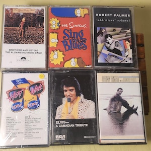 May include: A selection of vintage cassette tapes with album artwork. Titles include "Brothers and Sisters," "The Simpsons Sing the Blues," and "Elvis: A Canadian Tribute." Other tapes feature Robert Palmer and a "USA Wave" design.