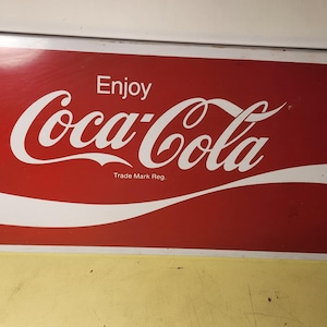 May include: A rectangular metal sign featuring a red background with the white text Enjoy Coca-Cola and the smaller text Trade Mark Reg. The sign includes a prominent white swoosh graphic across the bottom portion and two small mounting holes.