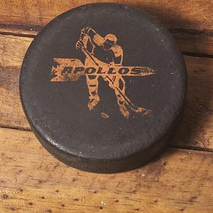 Op de afbeelding: Een zwarte hockeypuck met een oranje afbeelding van een hockeyspeler en het woord "APOLLOS". De puck is rond en ligt op een houten oppervlak. Een sportartikel.