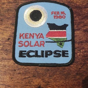Könnte beinhalten: Ein blauer gestickter Aufnäher mit schwarzem Rand. Der Aufnäher zeigt einen weißen Kreis mit einem schwarzen Kreis darin, eine Karte von Kenia mit den Farben der kenianischen Flagge sowie den Text "Kenya Solar Eclipse" und "Feb. 16, 1980".
