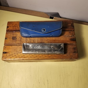 Könnte beinhalten: Eine Vintage-Holzkiste mit einem blauen Etui und einer silbernen Mundharmonika. Das blaue Etui hat einen Knopfverschluss. Die Mundharmonika ist silberfarben und trägt den Aufdruck "Echo". Die Holzkiste hat ein rustikales Aussehen.