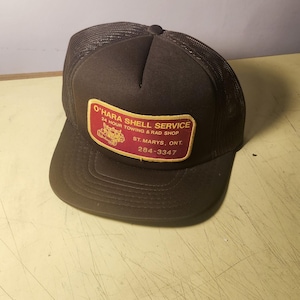 May include: A brown trucker cap with a mesh back and a solid front panel. A rectangular patch on the front features red text reading O'HARA SHELL SERVICE, 24 HOUR TOWING & RAD SHOP, ST. MARYS, ONT. and the phone number 284-3347 with a tow truck graphic.