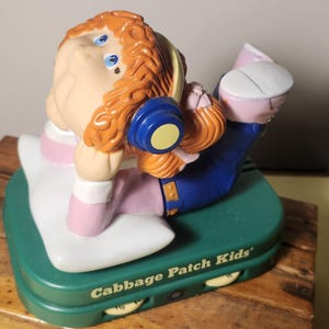 以下が含まれることがあります： ヨガのポーズをとるキャベツパッチキッズのフィギュア。青いヘッドフォンを着用しています。フィギュアは「Cabbage Patch Kids」の文字がプリントされた緑色の台座の上にあります。フィギュアは木製の表面にあります。