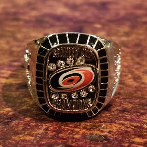 Puede incluir: Un anillo de plata con un logotipo de los Carolina Hurricanes en negro y rojo y las palabras "Stanley Cup Champions" en él.
