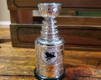 Pode incluir: Uma réplica prateada do troféu da Stanley Cup com um logotipo preto e branco dos San Jose Sharks no lado. O logotipo apresenta um tubarão com o texto "NHL" abaixo.