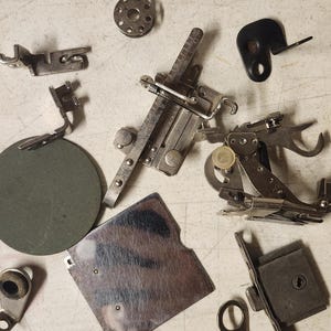 K&ouml;nnte beinhalten: Eine Ansammlung von Vintage-N&auml;hmaschinen-Teilen, darunter N&auml;hf&uuml;&szlig;e, Spulen und Zubeh&ouml;r, auf einer hellen Oberfl&auml;che. Die Metallkomponenten weisen verschiedene Formen und Gr&ouml;&szlig;en auf, mit silbernen und dunkleren Farbt&ouml;nen.