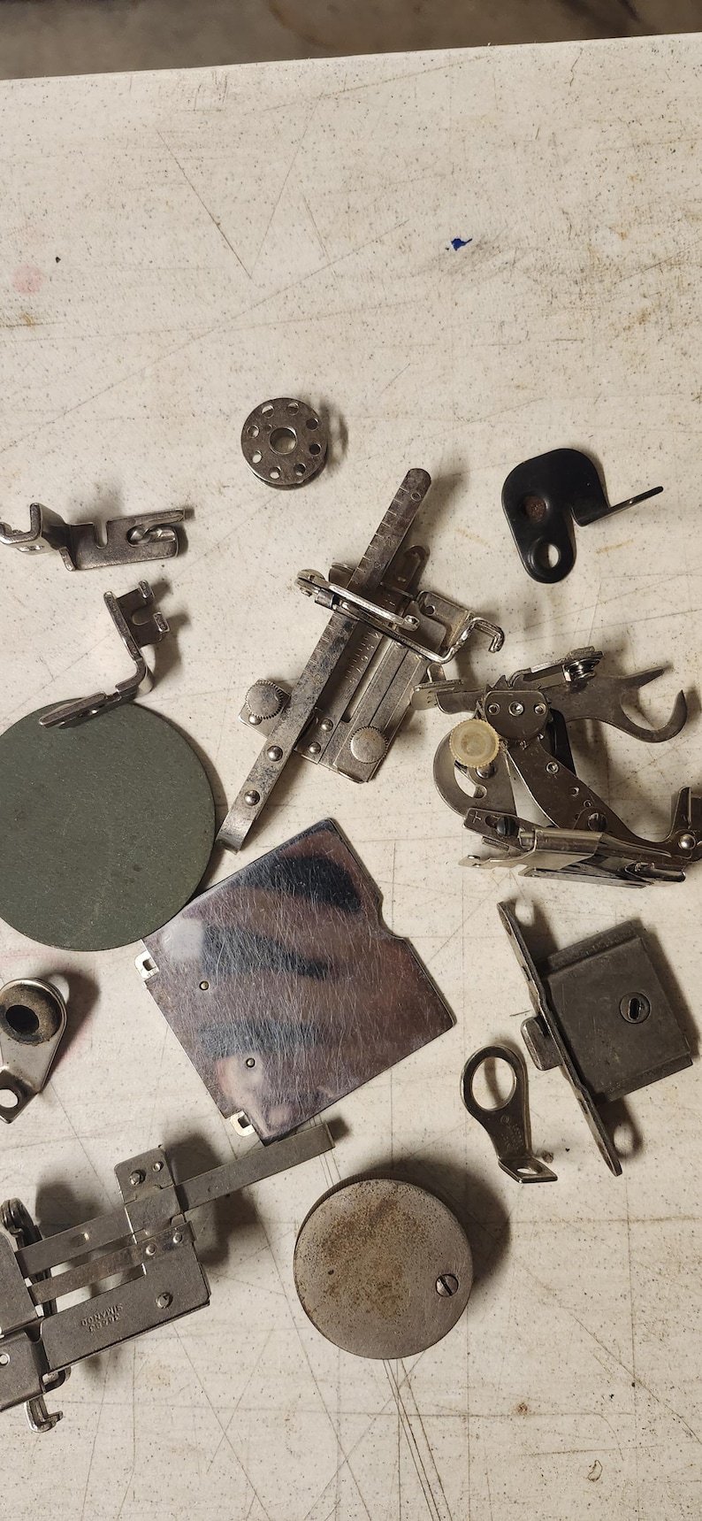 K&ouml;nnte beinhalten: Eine Ansammlung von Vintage-N&auml;hmaschinen-Teilen, darunter N&auml;hf&uuml;&szlig;e, Spulen und Zubeh&ouml;r, auf einer hellen Oberfl&auml;che. Die Metallkomponenten weisen verschiedene Formen und Gr&ouml;&szlig;en auf, mit silbernen und dunkleren Farbt&ouml;nen.
