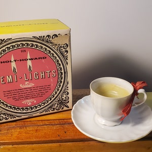 Puede incluir: Una caja amarilla y negra con el texto "Holt-Howard Demi-Lights 5125" en ella. La caja está sentada sobre una superficie de madera junto a una taza de té blanca con una cinta roja.