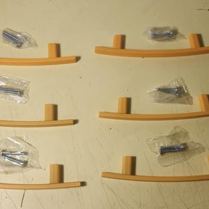 May include: Six wooden cabinet pulls with a curved design and two vertical posts. Each pull is a light orange color. Small bags of screws are also included.