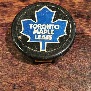 May include: A black hockey puck with a white Toronto Maple Leafs logo featuring a blue maple leaf.