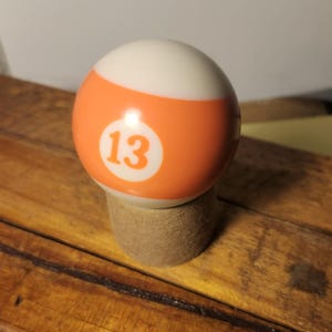 Peut inclure: Une boule de billard, blanche et orange, portant le numéro 13. Elle repose sur un petit support cylindrique, sur une surface en bois.