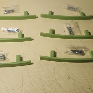 Könnte beinhalten: Sechs hellgrüne Schrankgriffe mit passenden Schrauben in einzelnen Plastiktüten. Die Griffe haben ein gebogenes Design mit zwei kleinen Pfosten an jedem Ende. Die Hardware ist neu und installationsbereit.