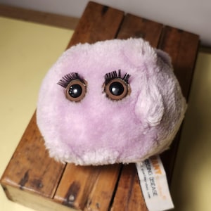 Pode incluir: Um brinquedo de pelúcia roxo com grandes olhos castanhos e cílios longos. O brinquedo tem a forma de uma esfera e uma textura felpuda.