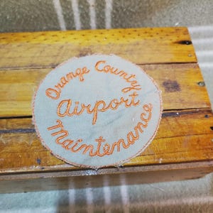 May include: A circular patch with the words "Orange County Airport Maintenance" embroidered in orange thread on a light blue background. The patch is set on a wooden surface.
