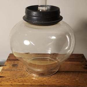 Puede incluir: Un tarro de vidrio transparente con una tapa de goma negra y un tubo de vidrio transparente que se extiende desde la tapa. El tarro está sentado sobre una superficie de madera.