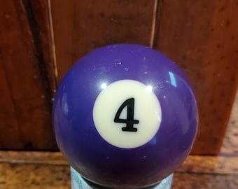 May include: A purple billiard ball with a white number 4 on it. The ball is sitting on a blue and white patterned base.