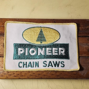May include: A rectangular embroidered patch with a white background and a yellow border. It features a green oval containing a stylized green pine tree, with the word PIONEER in white letters on a green rectangle and CHAIN SAWS below.