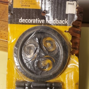 May include: Two black metal decorative holdbacks with a floral design. The holdbacks are packaged on a yellow card with the text "No. 1210/5 BLACK FINISH SPANISH decorative holdback PRICE".