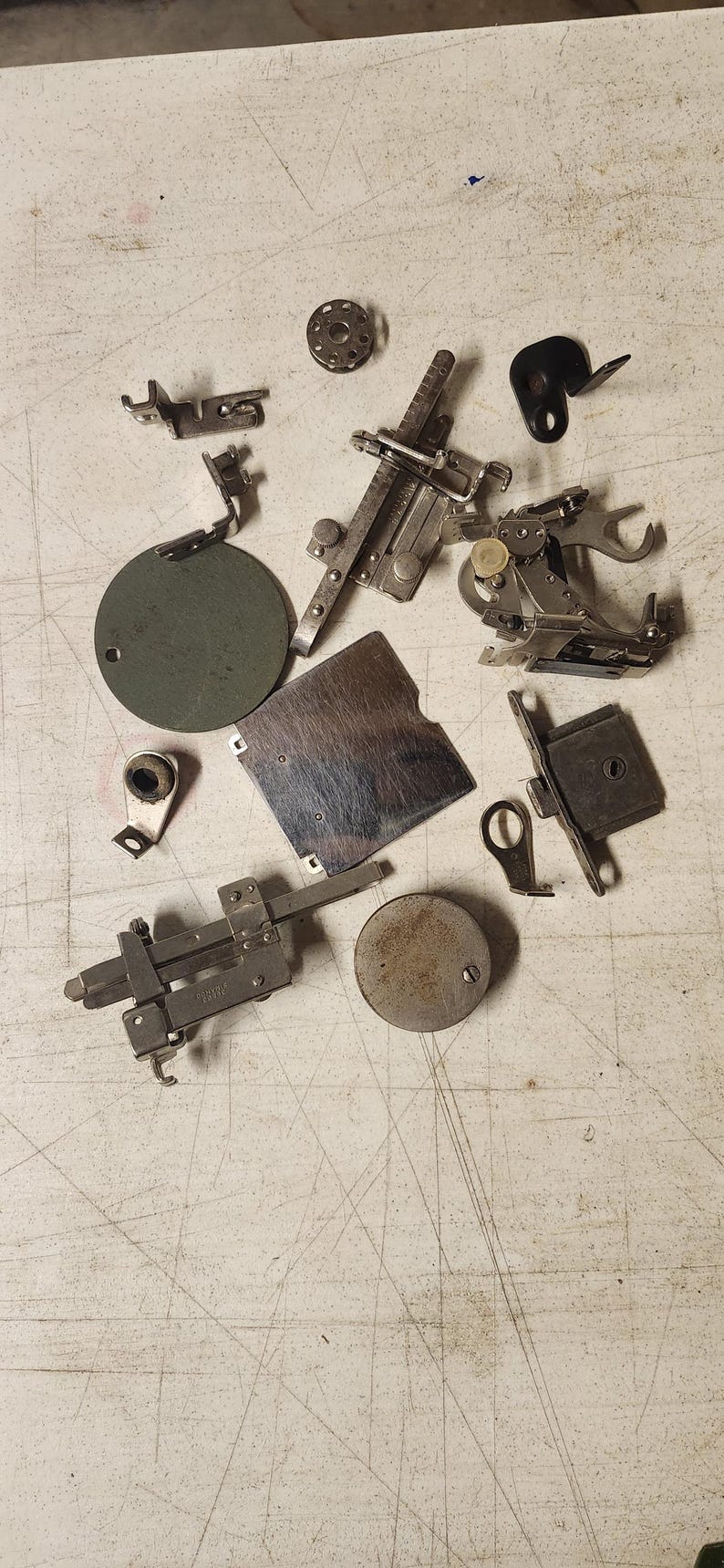 K&ouml;nnte beinhalten: Eine Ansammlung von Vintage-N&auml;hmaschinen-Teilen, darunter N&auml;hf&uuml;&szlig;e, Spulen und Platten, auf einer hellen Oberfl&auml;che. Die Metallkomponenten weisen Gebrauchsspuren auf und haben verschiedene Formen und Gr&ouml;&szlig;en.