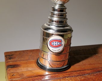 May include: A silver replica of the Stanley Cup trophy with a red, white and blue Montreal Canadiens logo on the side. The trophy is sitting on a wooden surface.
