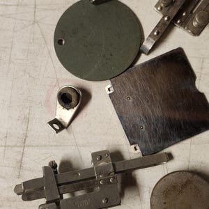 K&ouml;nnte beinhalten: Eine Auswahl an Vintage-N&auml;hmaschinen-Teilen, darunter N&auml;hf&uuml;&szlig;e, eine Spule und ein Messger&auml;t. Die Metallteile sind auf einer hellen Oberfl&auml;che angeordnet. Auf dem Messger&auml;t steht "DONVIS SIMANCO 36403".
