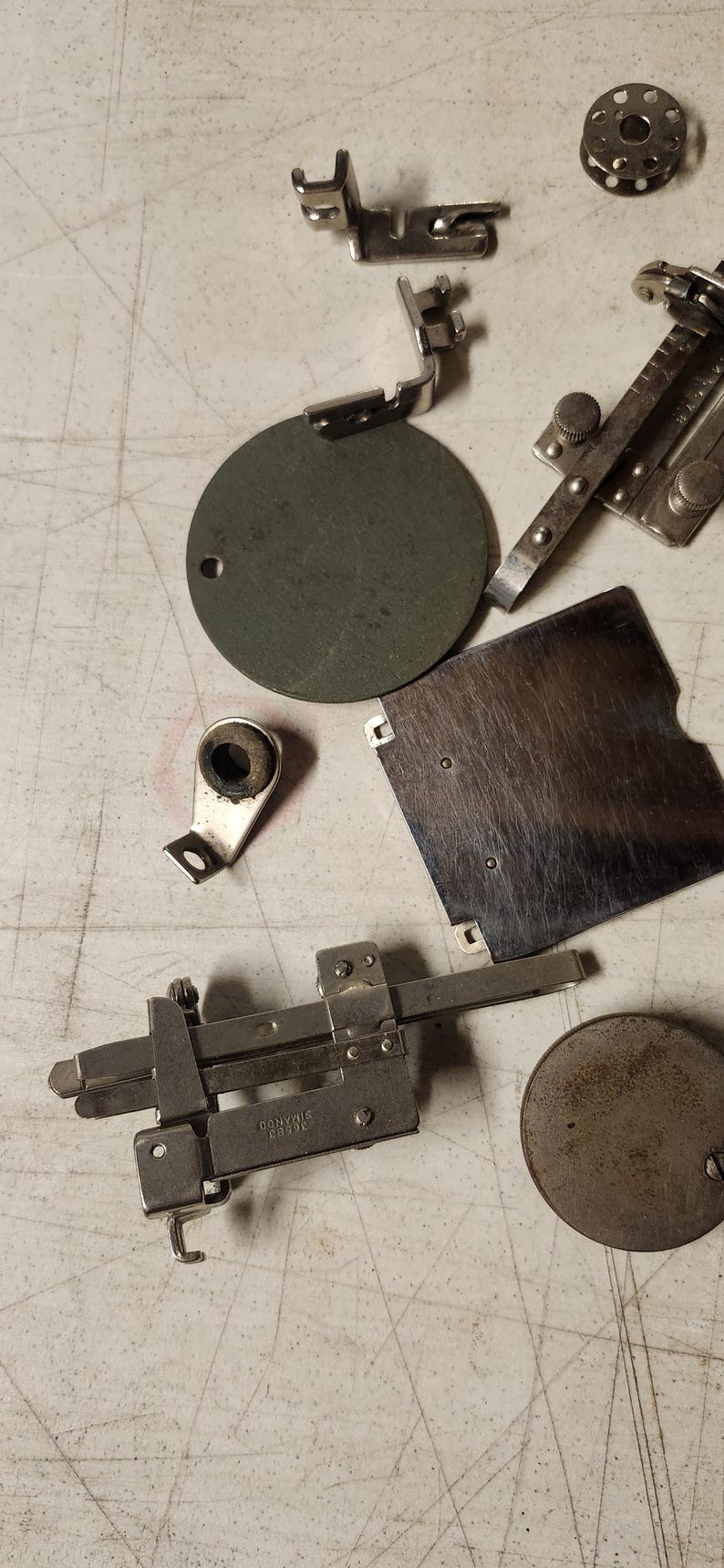 K&ouml;nnte beinhalten: Eine Auswahl an Vintage-N&auml;hmaschinen-Teilen, darunter N&auml;hf&uuml;&szlig;e, eine Spule und ein Messger&auml;t. Die Metallteile sind auf einer hellen Oberfl&auml;che angeordnet. Auf dem Messger&auml;t steht "DONVIS SIMANCO 36403".