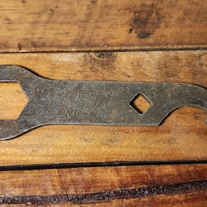 May include: A weathered metal multi-tool wrench, lying on a wooden surface. The wrench features a hexagonal opening, a diamond-shaped hole, and a curved end. The text "ENGLAND" is visible on the wrench.