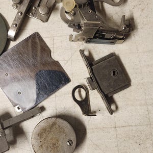 K&ouml;nnte beinhalten: Eine Ansammlung von Vintage-N&auml;hmaschinen-Teilen, darunter N&auml;hf&uuml;&szlig;e, Spulen und Messger&auml;te, liegt auf einer hellen Oberfl&auml;che. Die Metallkomponenten weisen Gebrauchsspuren auf, was darauf hindeutet, dass sie antik oder stark genutzt wurden.