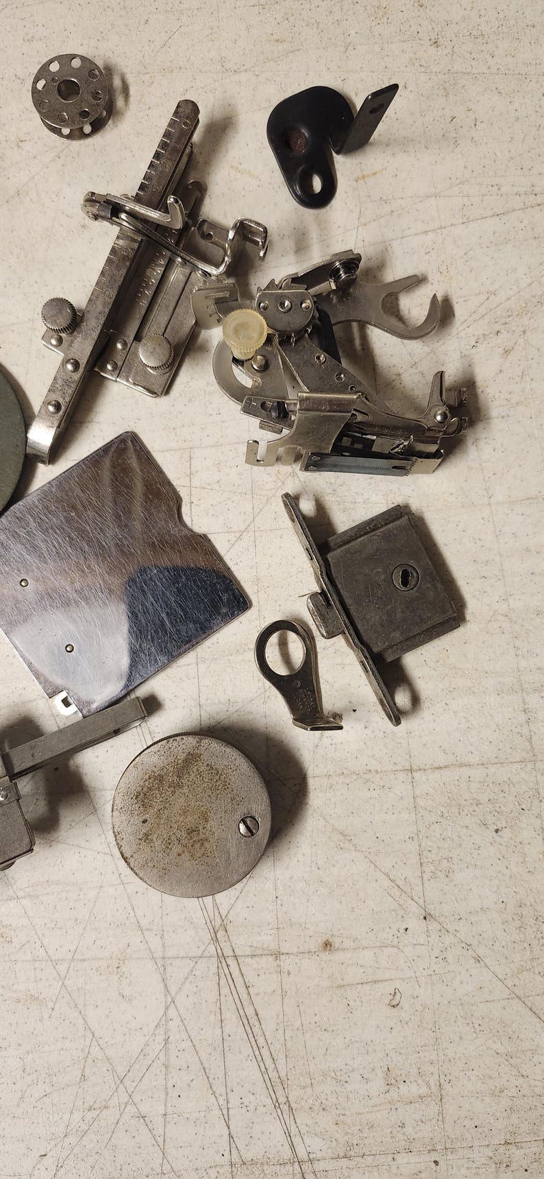 K&ouml;nnte beinhalten: Eine Ansammlung von Vintage-N&auml;hmaschinen-Teilen, darunter N&auml;hf&uuml;&szlig;e, Spulen und Messger&auml;te, liegt auf einer hellen Oberfl&auml;che. Die Metallkomponenten weisen Gebrauchsspuren auf, was darauf hindeutet, dass sie antik oder stark genutzt wurden.