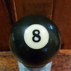 以下が含まれることがあります： 数字の8が描かれた白い円が付いた黒いビリヤードボール。淡い青と白の模様の小さなカップの上に置かれています。背景は濃い木製の表面です。