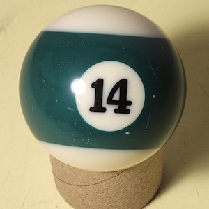 Peut inclure: Une boule de billard, avec une partie supérieure blanche et une section inférieure vert d'eau, portant le numéro 14 en noir. Elle repose sur une petite base cylindrique en carton. Un accessoire de jeu classique.