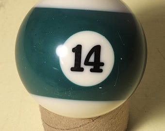 May include: A billiard ball, featuring a white top and a teal green lower section, with the number 14 in black. The ball is resting on a small, cylindrical cardboard base. A classic game accessory.