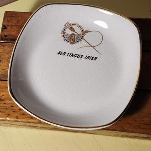 Peut inclure: Un plat carré en céramique blanche avec un bord doré. Le plat présente un motif doré et coloré d'une harpe et d'une aiguille avec le texte "AER LINGUS - IRISH" en dessous.