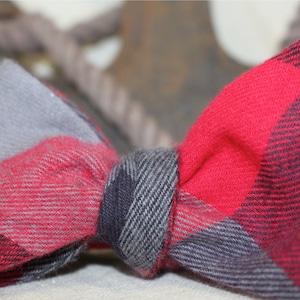 May include: A red, gray, and black plaid bow tie. The bow tie is made of a soft, woven fabric.