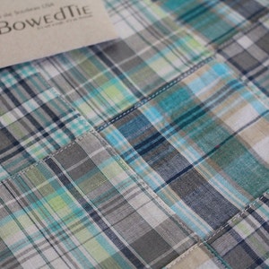 May include: Close-up of a patchwork fabric with a plaid pattern in shades of gray, teal, and white. The fabric is stitched together in a grid-like design. A tag with the text "The Bowed Tie" is visible.