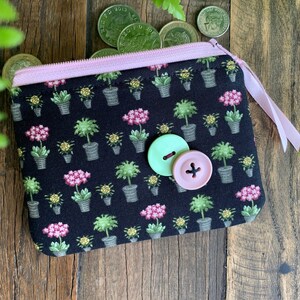 May include: A black zippered pouch with a pink zipper and pink trim. The pouch has a floral print with pink flowers and green leaves. Two buttons are attached to the front of the pouch, one green and one pink.