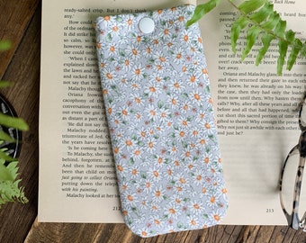 May include: A glasses case with a white button closure, featuring a white and orange floral print. The case is lying on a book with text.