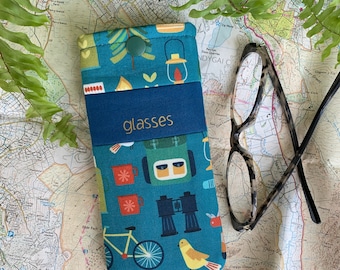 May include: A blue and green glasses case with a camping theme. The case has a zipper closure and features a variety of camping-related images, including a tent, a compass, binoculars, and a campfire. The text "glasses" is printed on the front of the case.