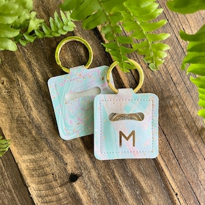 May include: Two keyrings with a light blue and pink marbled pattern. Each keyring has a gold-coloured ring and a square pouch with a cutout. One pouch has the letter "M" printed on it.