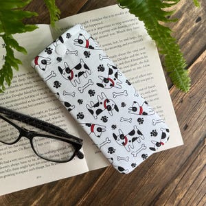 May include: White glasses case featuring a pattern of black and white dogs, bones, and paw prints. The dogs have red collar details and the case has a button closure. The case is on an open book.