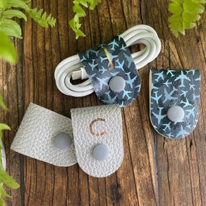 May include: Three cable organizers, two are gray with a button closure and one is blue and black with a plane pattern. The gray organizers have a letter C embossed on them.
