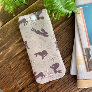 May include: A fabric glasses case with a button closure. The case has a repeating pattern of running brown rabbits, floral elements, and white dots on a light beige background. The rectangular case is designed to hold eyeglasses.