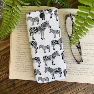 May include: A white and black zebra print glasses case with a black button closure. The case is made of fabric and has a white background with black zebra stripes.