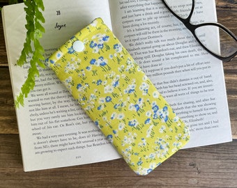 May include: A yellow and blue floral fabric eyeglass case with a white button closure. The case is lying on a book with text.