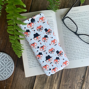 May include: A white and orange glasses case with a repeating pattern of foxes wearing glasses, hats, and mustaches. The case has a small button closure.