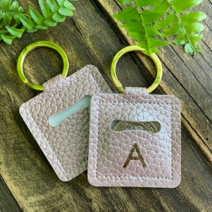 May include: Two light pink leather keyrings with gold key rings. One keyring has a coin pocket and the letter "A" embossed on the front. The other keyring has a coin pocket.