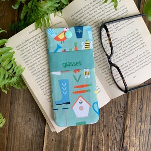 May include: A light blue glasses case with a colorful garden print featuring a bird, a house, boots, carrots, and bees. The case has a green flap with the word "glasses" printed on it.