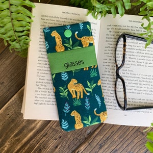 Peut inclure: Un étui à lunettes en tissu vert et noir avec un imprimé léopard et des feuilles vertes. L'étui a un rabat avec une languette verte qui dit "glasses".